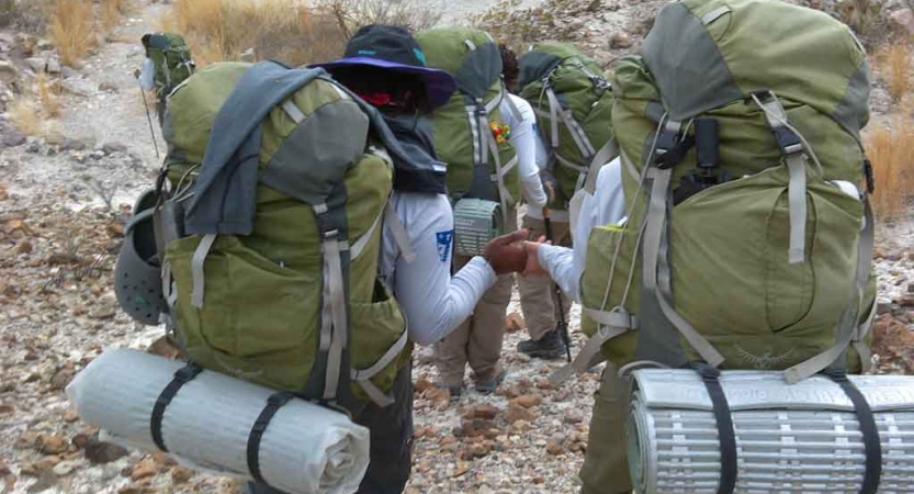 a group of students carrying backpacks descend over a rocky landscape. two of the students hold hands as they hike. 